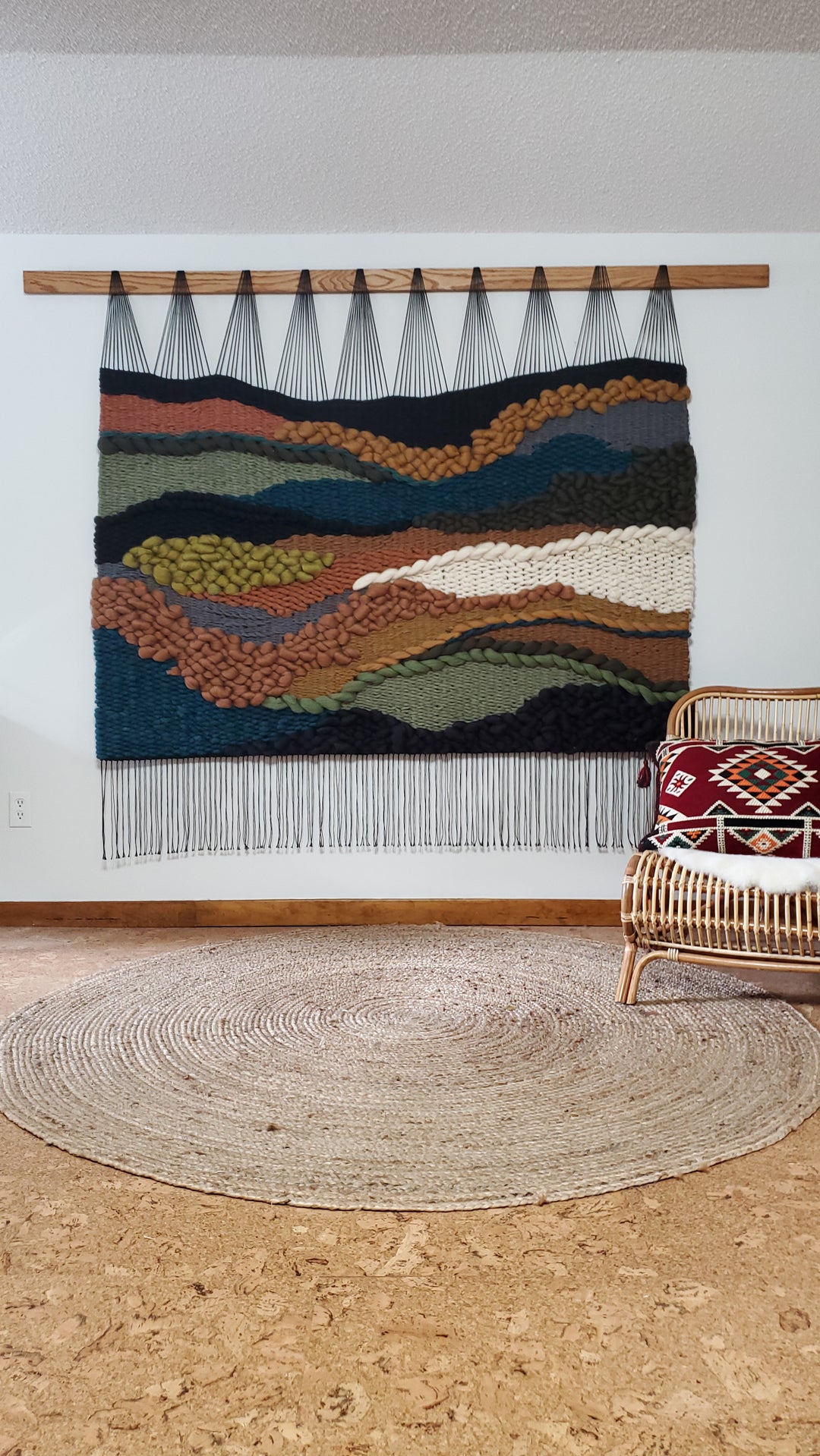 Woven wall art with a mountain landscape design in a room with a woven rug and wicker chair.