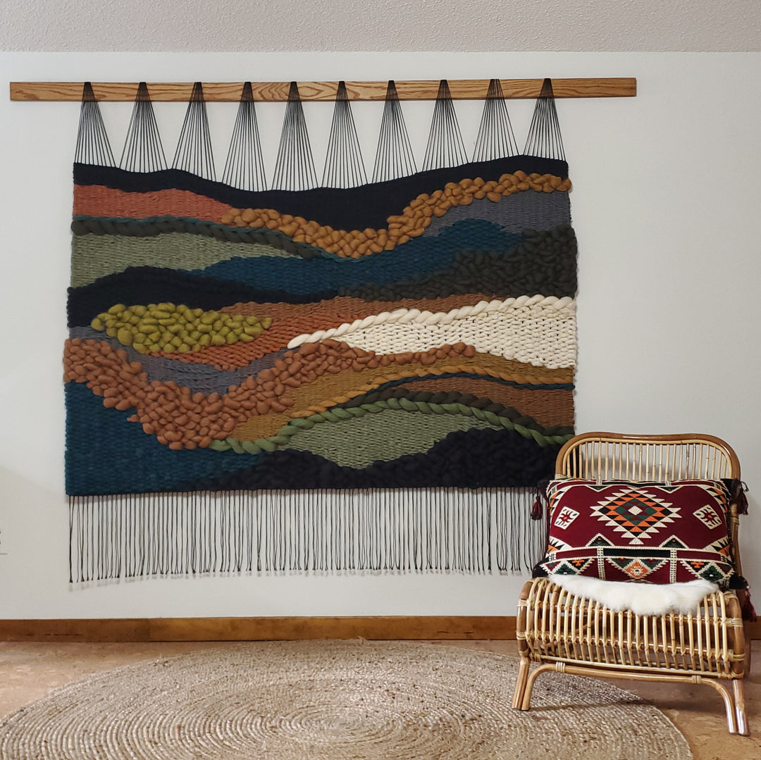 Woven wall art with a textured landscape design in a room with a chair and rug.