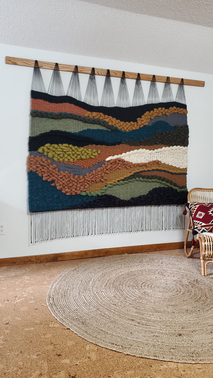 Colorful woven wall art hanging on a white wall with a round beige rug on the floor.